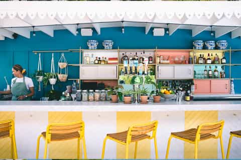 Colorful outdoor bar counter with teal back wall, yellow chairs, and shelves of spirits and plants
