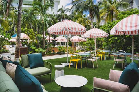 Tropical garden lawn with striped pink and white umbrellas, colorful chairs, and palm trees