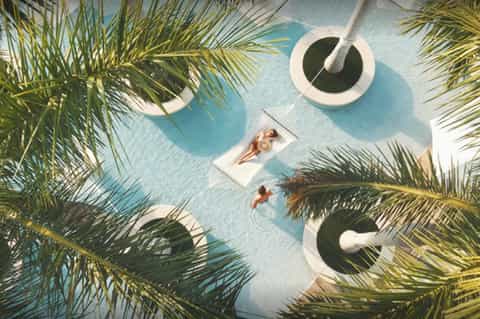 Aerial view of light blue pool with palm trees, white float raft, and sunbathing guests