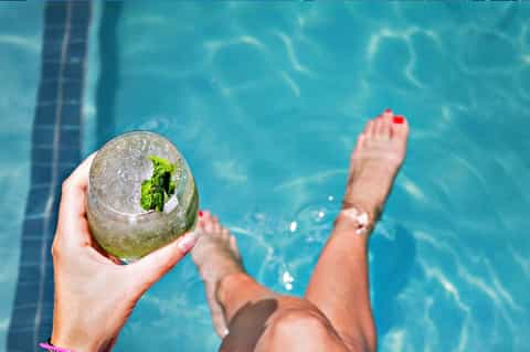 Person holding mojito cocktail by pool with feet relaxing in water