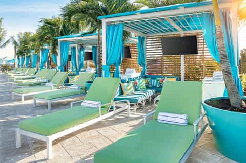 Beachside cabanas with turquoise pergolas, green loungers, and palm trees under clear sky