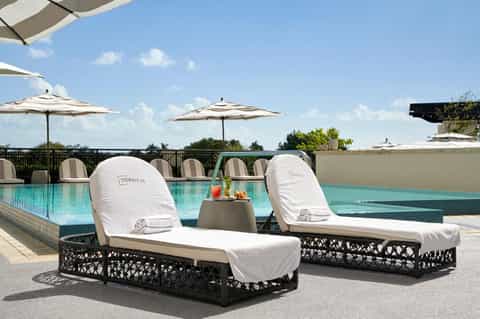 Two lounge chairs with white cushions beside turquoise pool under umbrellas