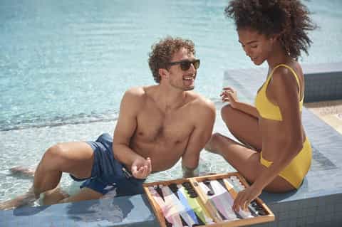 Couple in swimwear sitting by pool edge holding color swatches, enjoying tropical resort atmosphere
