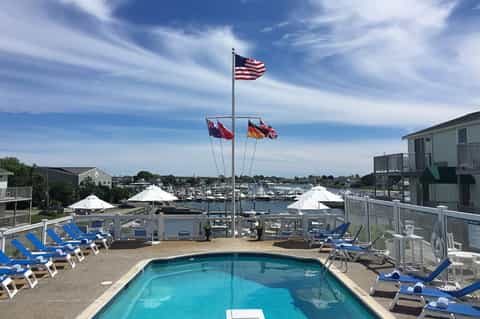 Waterfront resort pool with blue loungers, white umbrellas, and sailboats visible in harbor beyond