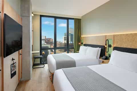 Modern hotel room with two queen beds, floor-to-ceiling windows overlooking city skyline, and contemporary furnishings