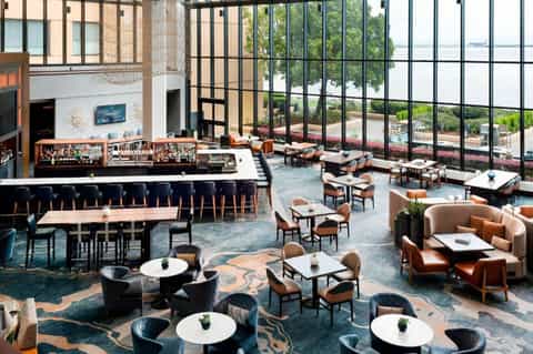 Modern hotel lobby with high ceilings, bar seating, lounge areas, and floor-to-ceiling windows overlooking water