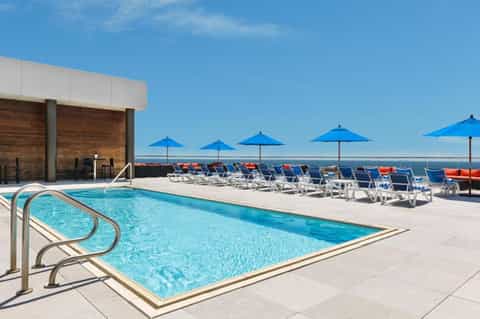 Resort pool deck with blue umbrellas, white lounge chairs, and ocean view on clear sunny day