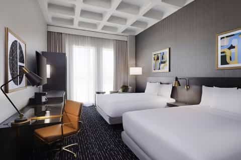 Modern hotel room with dark gray accent wall, two beds, workspace with monitor, coffered white ceiling, and contemporary furnishings