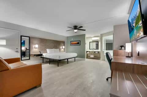 Spacious hotel suite with king bed, living area with tan leather chair, desk workspace, and ensuite bathroom