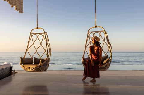 Woman in rust-colored dress standing between two woven hanging egg chairs on a beach at golden hour