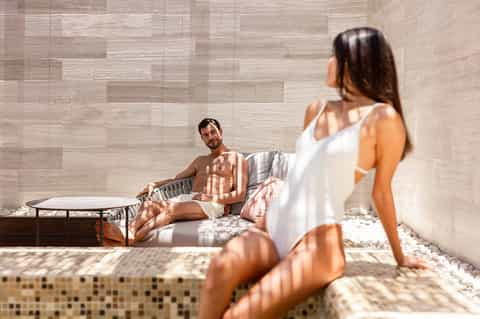Spa relaxation area with couple on lounger, neutral tile walls, and natural sunlight