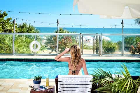 Woman in bikini relaxing by outdoor pool overlooking ocean with palm trees and beach cabanas