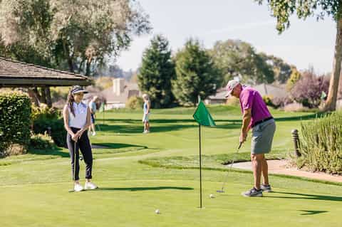 Golfers on manicured fairway with flag pole, trees, and clubhouse shelter on sunny day