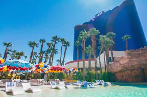 Resort pool with colorful umbrellas, lounge chairs, palm trees, and large blue hotel building with water slide