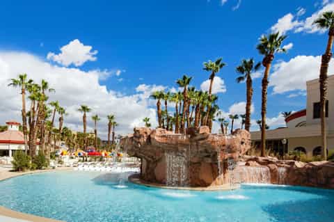 Resort pool with rock waterfall feature, palm trees, lounge chairs, and colorful umbrellas under blue sky