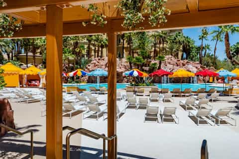 Poolside terrace with colorful umbrellas, lounge chairs, palm trees, and covered pergola structure