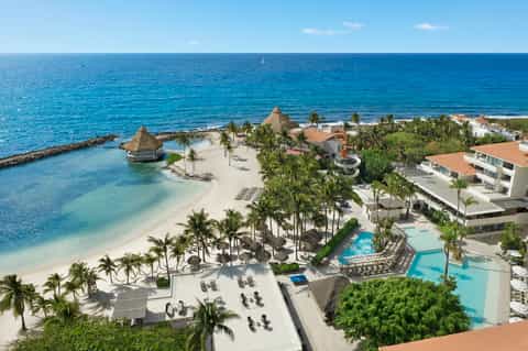 Aerial view of beachfront resort with pools, palm trees, thatched cabanas, and turquoise ocean water