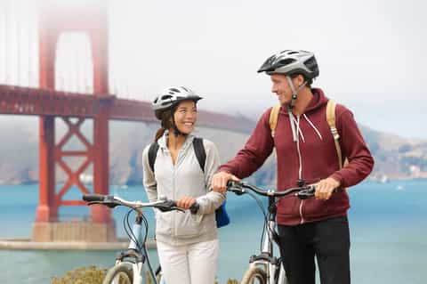 Two cyclists with helmets posing with bikes in front of Golden Gate Bridge and bay