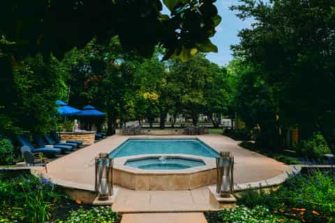 Resort pool with hot tub, loungers, blue umbrellas, and lush trees surrounding patio area
