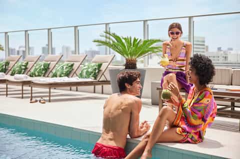 Woman and man enjoying drinks by a rooftop pool with city skyline views, lounge chairs, and palm plants