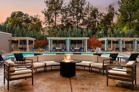Outdoor fire pit lounge with curved beige seating, turquoise pool, and covered cabanas under evening sky