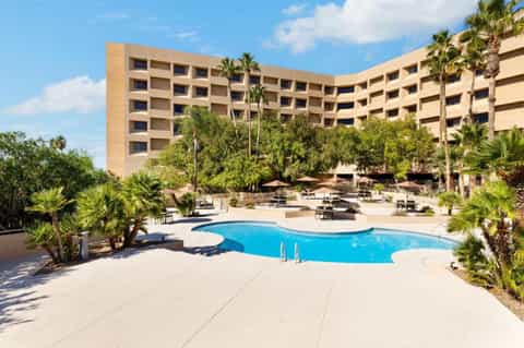 Resort pool with curved blue water, multi-story tan building, and palm trees