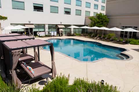 Resort pool area with cabanas, white umbrellas, lounge chairs, and multi-story building backdrop