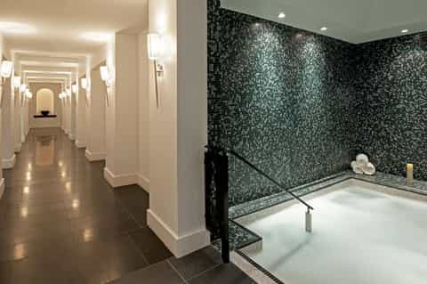 Luxury spa hallway with cream walls and sconces leading to a sunken soaking tub with black mosaic tile walls