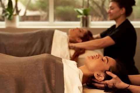 Spa therapists performing head and neck massages on clients in a bright, relaxing wellness room with plants