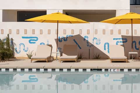 Modern resort pool deck with yellow umbrellas, lounge chairs, and blue wall art under bright sunlight