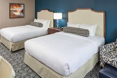 Hotel room with two queen beds, cream upholstered headboards, blue accent wall, and bedside lamp
