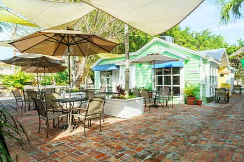 Outdoor patio area with umbrellas, dining tables, green cottage building, and tropical landscaping on brick walkway