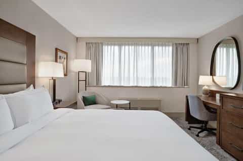 Spacious hotel bedroom with queen bed, work desk, armchair, and large windows with sheer curtains