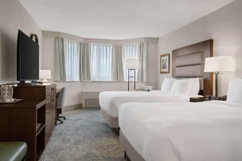 Hotel bedroom with two queen beds, wood furniture, and bay windows with curtains