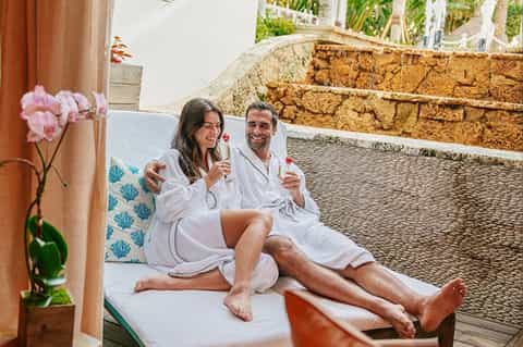 Couple in white robes toasting with champagne glasses on a terrace lounge chair