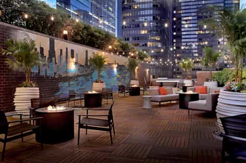 Rooftop terrace at dusk with fire pits, seating areas, mural backdrop, and city skyline views