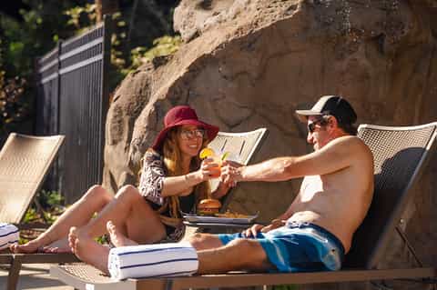 Couple sharing appetizers by a pool, seated in lounge chairs against a large rock formation