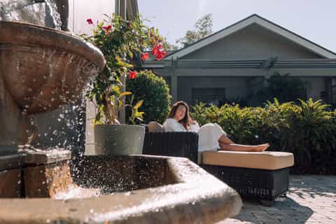Woman relaxing in wicker chair by courtyard fountain with flowering plants and modern home
