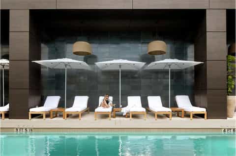 Luxury poolside lounge area with white umbrellas, wooden loungers, and glass wall backdrop