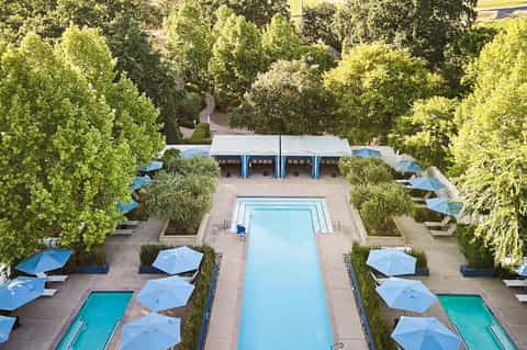 Aerial view of resort pool surrounded by blue umbrellas, loungers, lush trees, and cabana structures