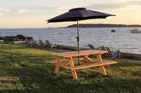 Waterfront picnic table with black umbrella on grass overlooking calm bay waters and distant shoreline