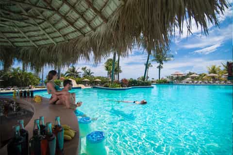 Resort guests relaxing by tropical lagoon pool with thatched roof shelter and palm trees