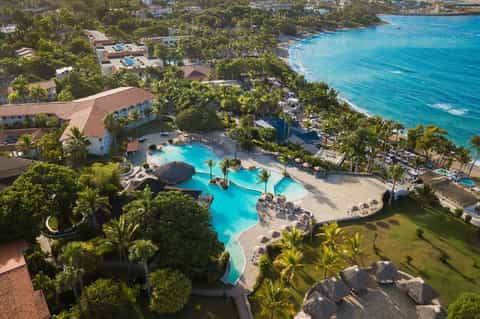 Aerial view of beachfront resort with curved pool, tropical grounds, and ocean