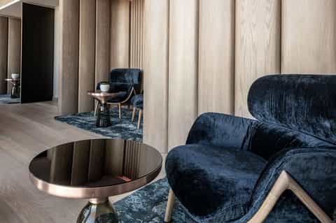 Contemporary lounge interior with light wood paneling, dark blue velvet armchairs, round black metal table, and neutral area rugs