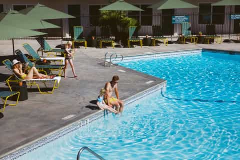Resort pool deck with lounging guests, yellow umbrellas, green shade structures, and clear blue water