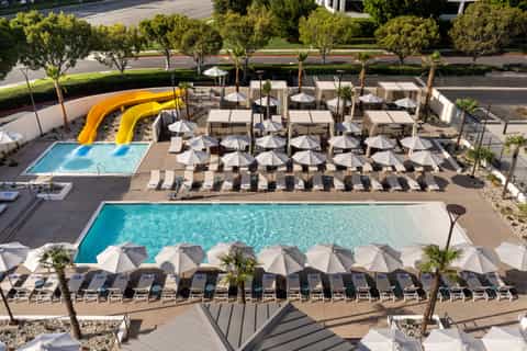 Aerial view of hotel pool deck with multiple pools, yellow water slides, white umbrellas, and palm trees