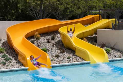 Two children sliding down bright yellow water slides into a resort pool with landscaped grounds