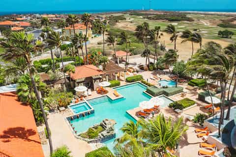 Aerial view of tropical resort with turquoise pools, palm trees, and orange-roofed buildings overlooking coastline