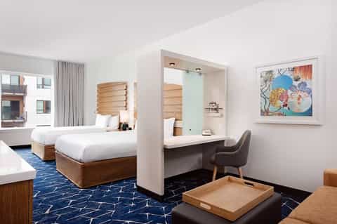 Modern hotel room with two queen beds, wooden headboards, desk with chair, artwork on wall, blue patterned carpet