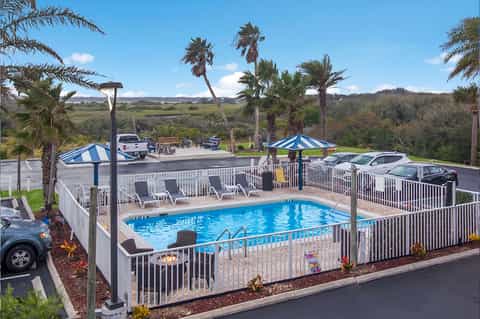 Hotel pool surrounded by white fence, blue umbrellas, palm trees, and hillside landscape view
